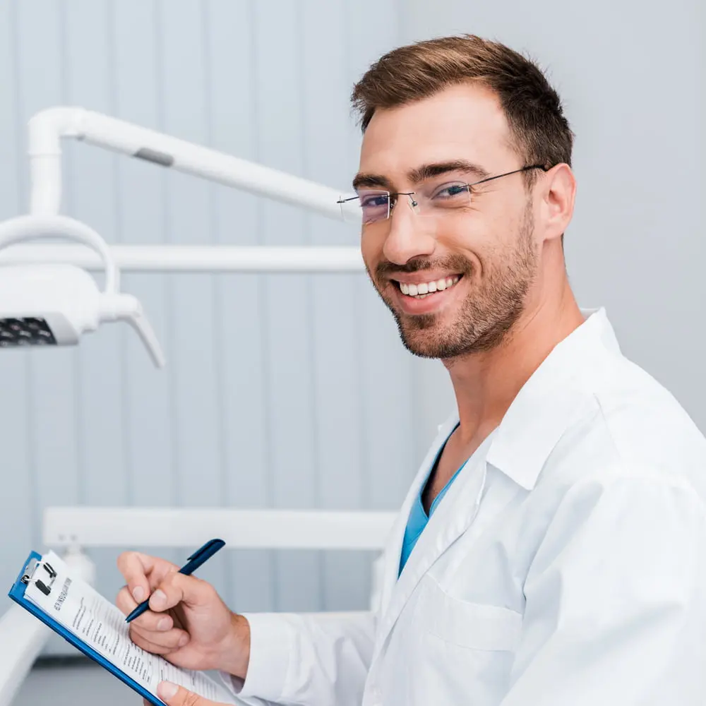 A male dentist in a white coat smiles while holding a clipboard and pen in a dental office in Austin