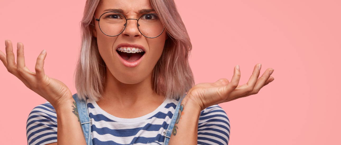 A person with glasses and braces looks surprised, with hands raised. They have shoulder-length light hair and wear a striped shirt against a pink background.