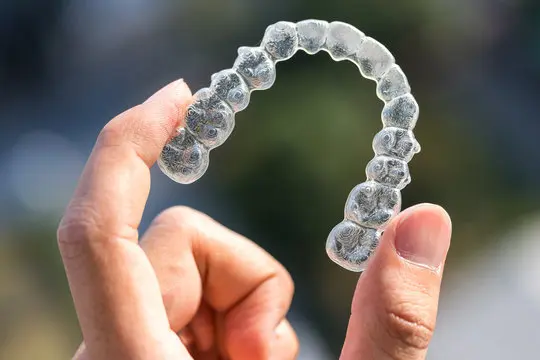 A hand from Best Orthodontics delicately holds a clear dental aligner against a softly blurred background.
