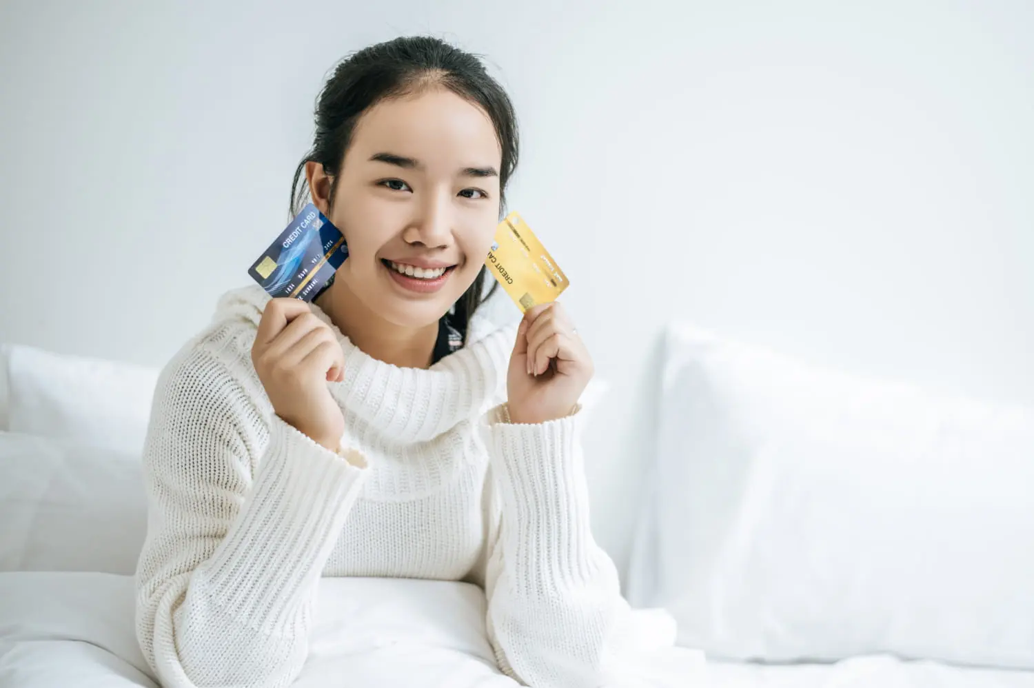 Seated on a bed with white sheets, a person in a white sweater beams while holding two credit cards, one blue and one yellow. As if unveiling their best orthodontics-approved smile, they radiate confidence and delight.