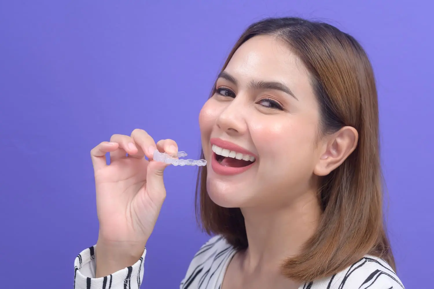 A person with medium-length hair smiles brightly while holding a clear dental aligner near their mouth against a purple background, showcasing the best orthodontics in action.