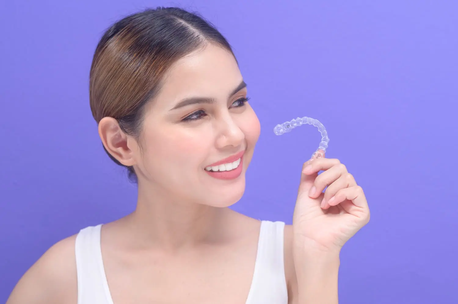 Person smiling while holding a transparent dental aligner against a purple background, showcasing the best orthodontics in modern dental care.