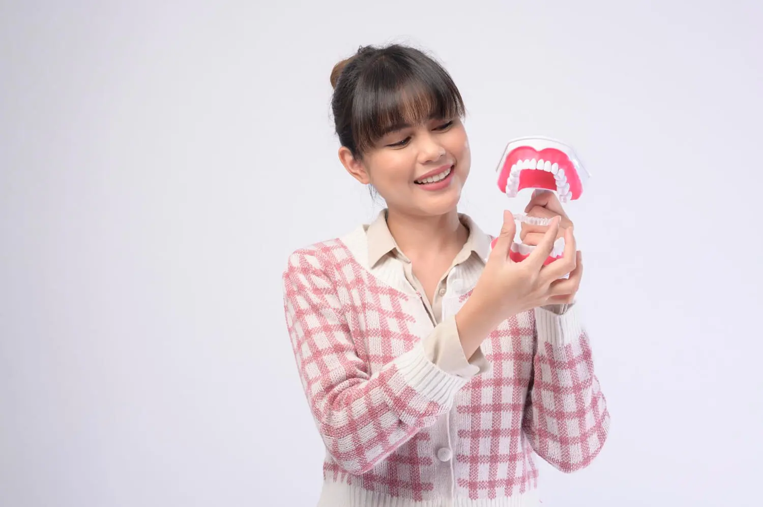 A woman with a checked sweater smiles warmly as she holds a dental model displaying teeth and gums, showcasing the best orthodontics against a plain background.