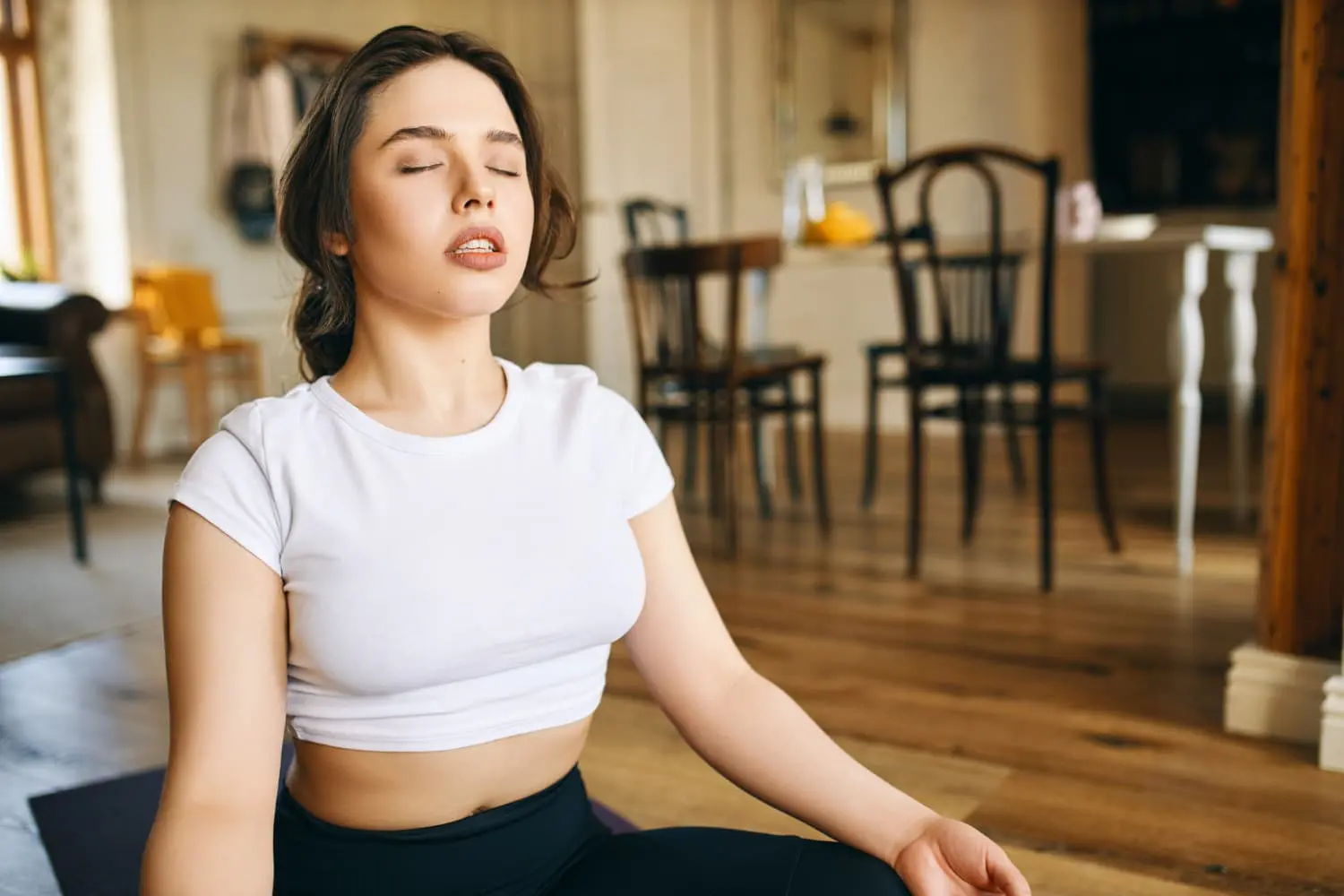 In a serene indoor setting, a woman in a white t-shirt and black pants sits cross-legged with eyes closed, radiating calmness. Just as the best orthodontics align smiles, her tranquil pose harmonizes body and mind.