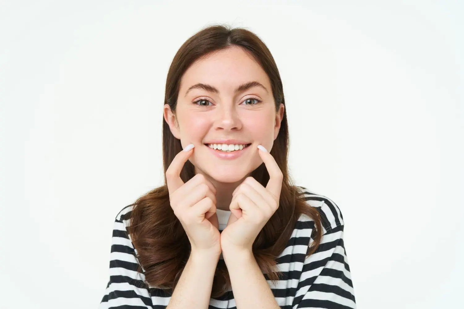 A smiling woman in a striped shirt points to her cheeks against a plain background, radiating confidence after her treatment