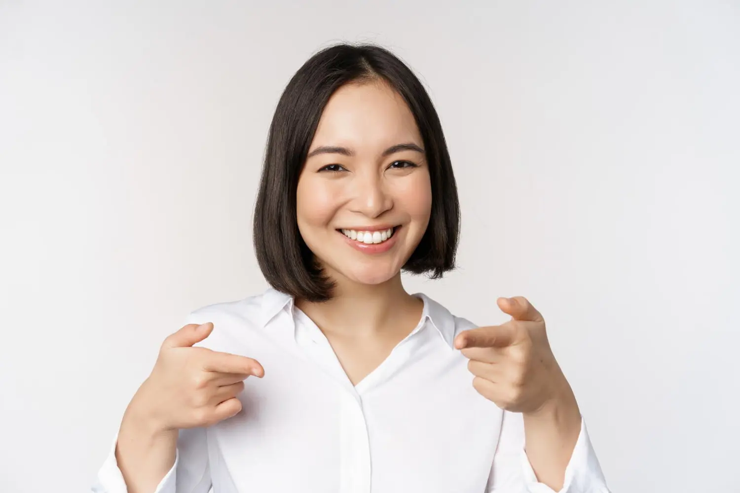 A smiling person with short dark hair, dressed in a white shirt, enthusiastically points with both index fingers against a plain background, embodying the confidence you get with Best Orthodontics.