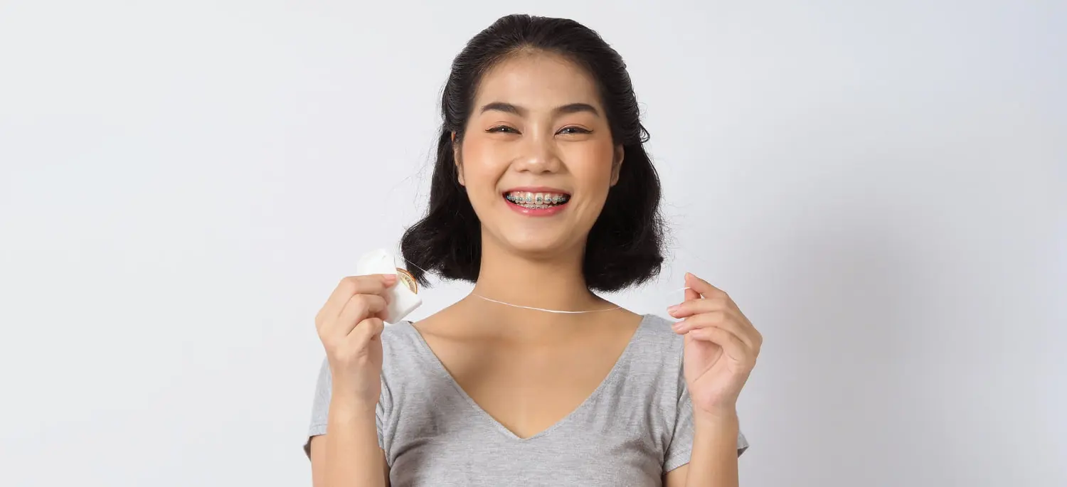 A beaming person shows off their braces, holding dental floss against a plain background, celebrating their commitment to a perfect smile in Houston