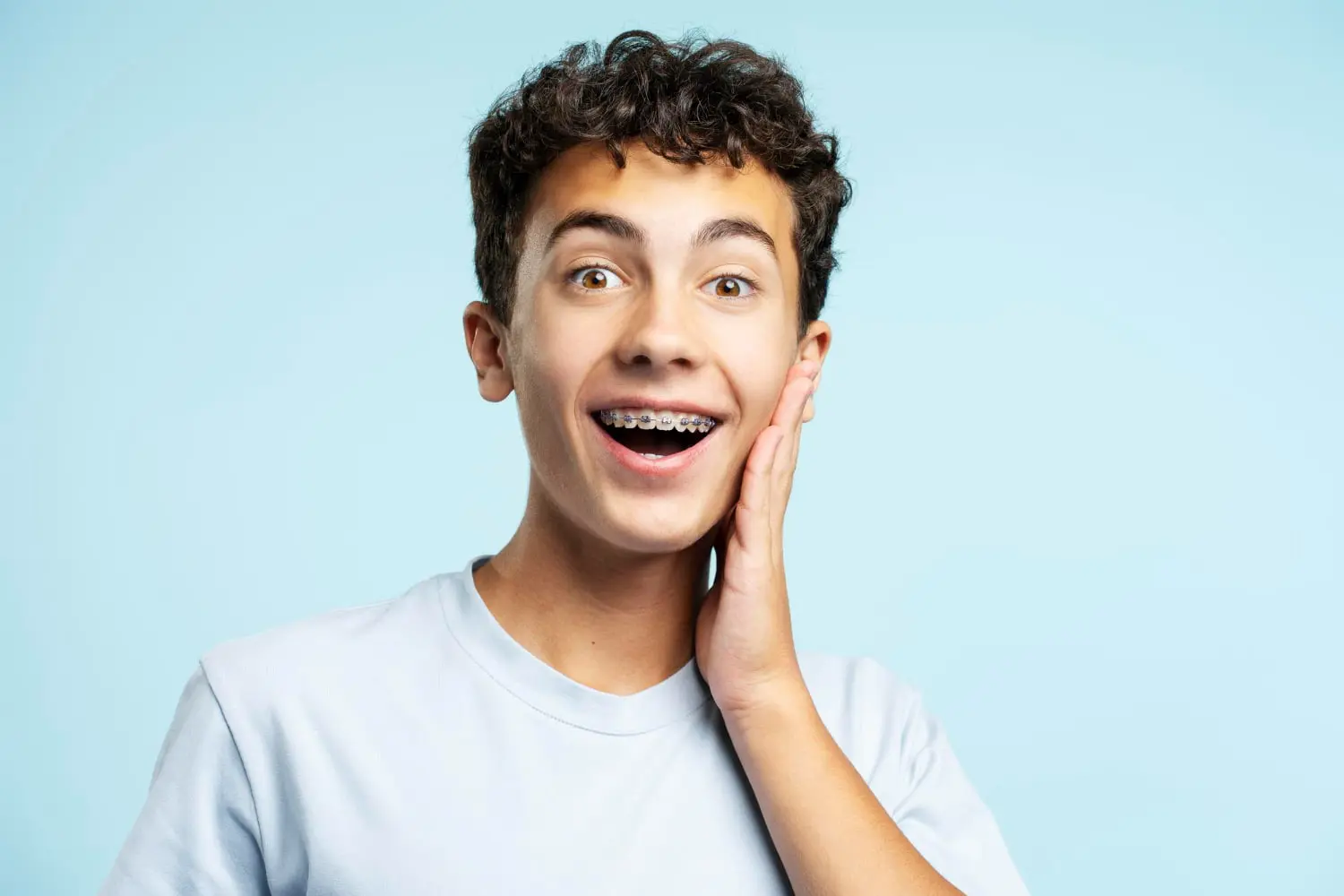 A teenage boy with curly hair and braces, courtesy of Best Orthodontics, smiles with surprise, holding his cheek against a light blue background.