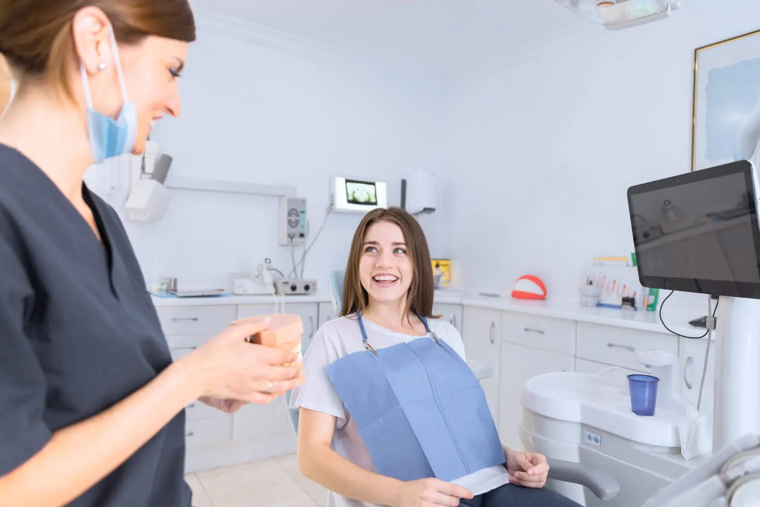 A woman sitting in a dental chair beams at a dental professional holding a cup, enjoying the comfort of the best orthodontics care. The room is equipped with advanced dental tools and technology.
