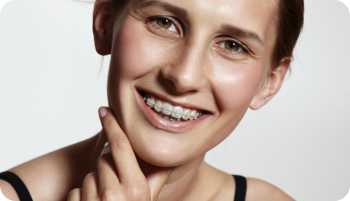 Woman smiling, wearing braces, with a hand touching her chin against a light background.