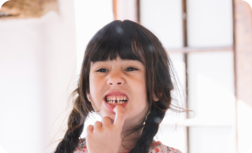 Child with dark hair and braided pigtails smiles, showing a gap where a tooth is missing, in a brightly lit room.