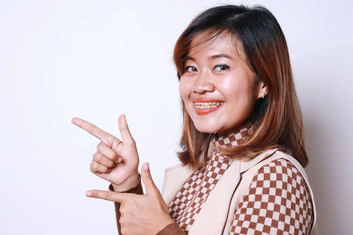 A woman with braces beams and points to the side, showcasing her stylish checkered brown turtleneck and beige vest. Her confident smile suggests she might know where to find the best orthodontics in town.