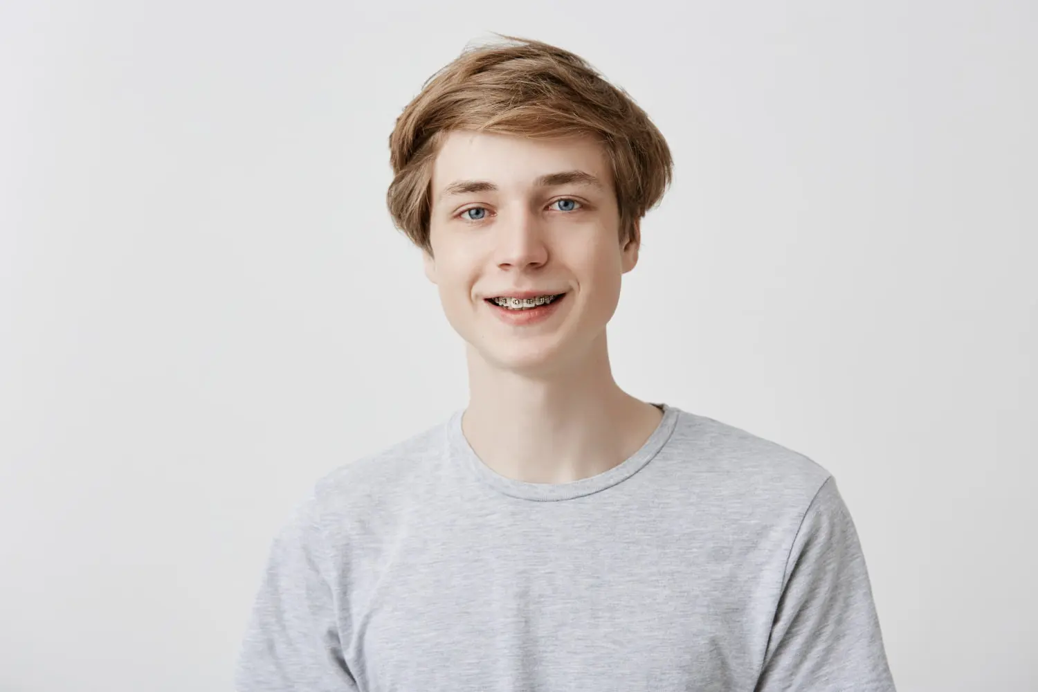 A young person with sparkling braces beams a radiant smile, wearing a gray T-shirt against a plain white background in Austin