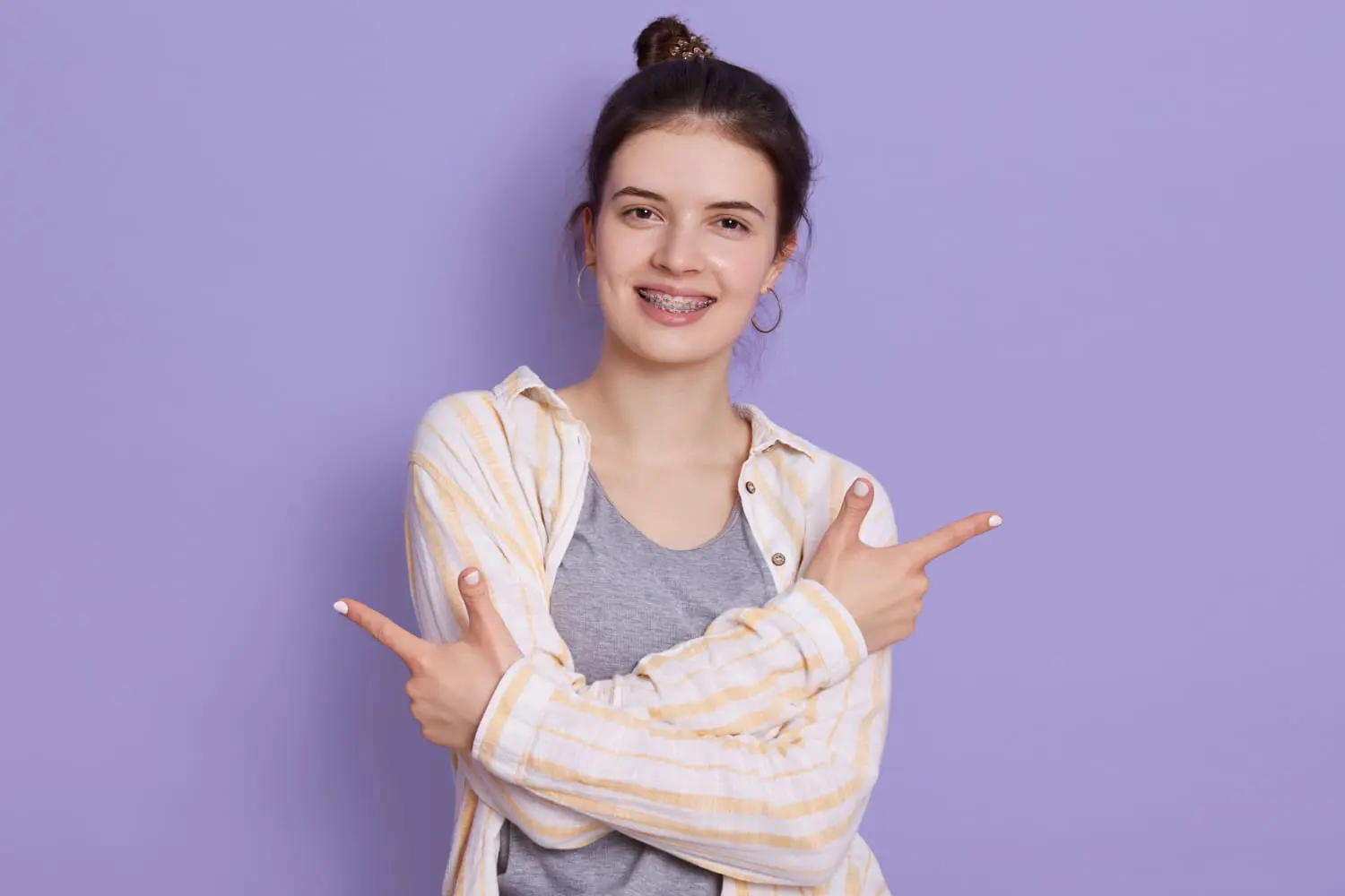 Woman with braces smiling, wearing a striped shirt over a gray top, points with both hands against a purple background, showcasing the results of Best Orthodontics.