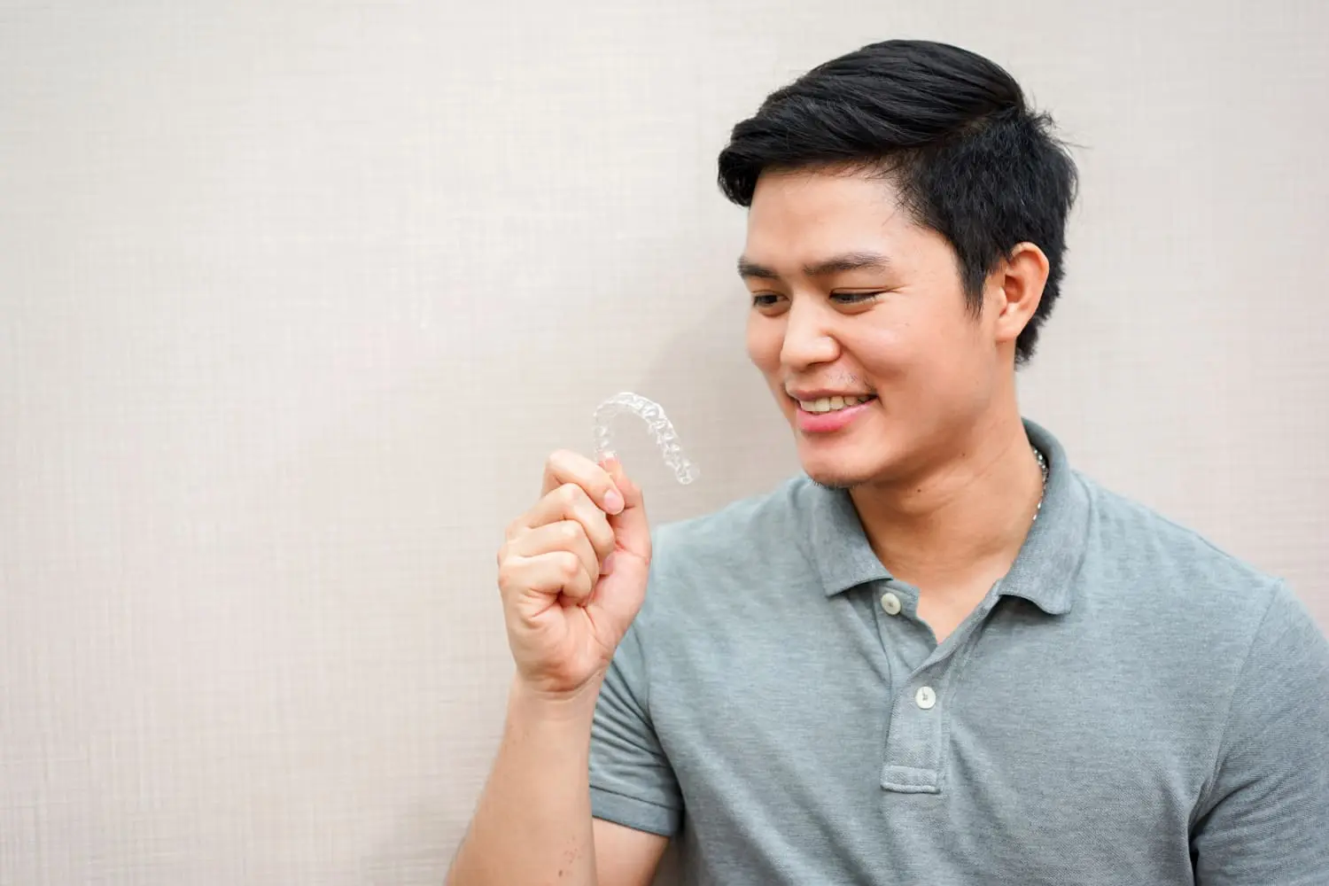A man in a gray polo shirt, showcasing the best orthodontics with a clear dental aligner, smiles warmly against a light-colored background.