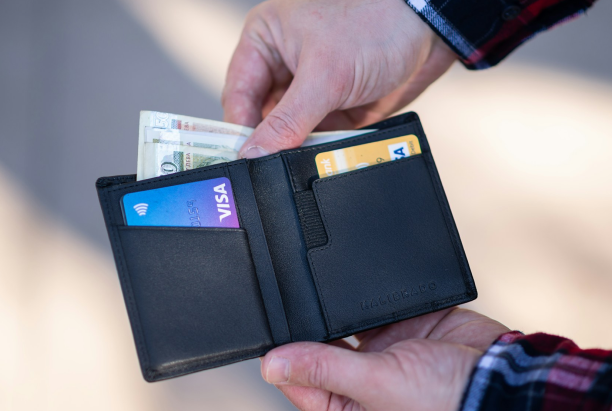A person holding a black wallet with visible cash and two credit cards.