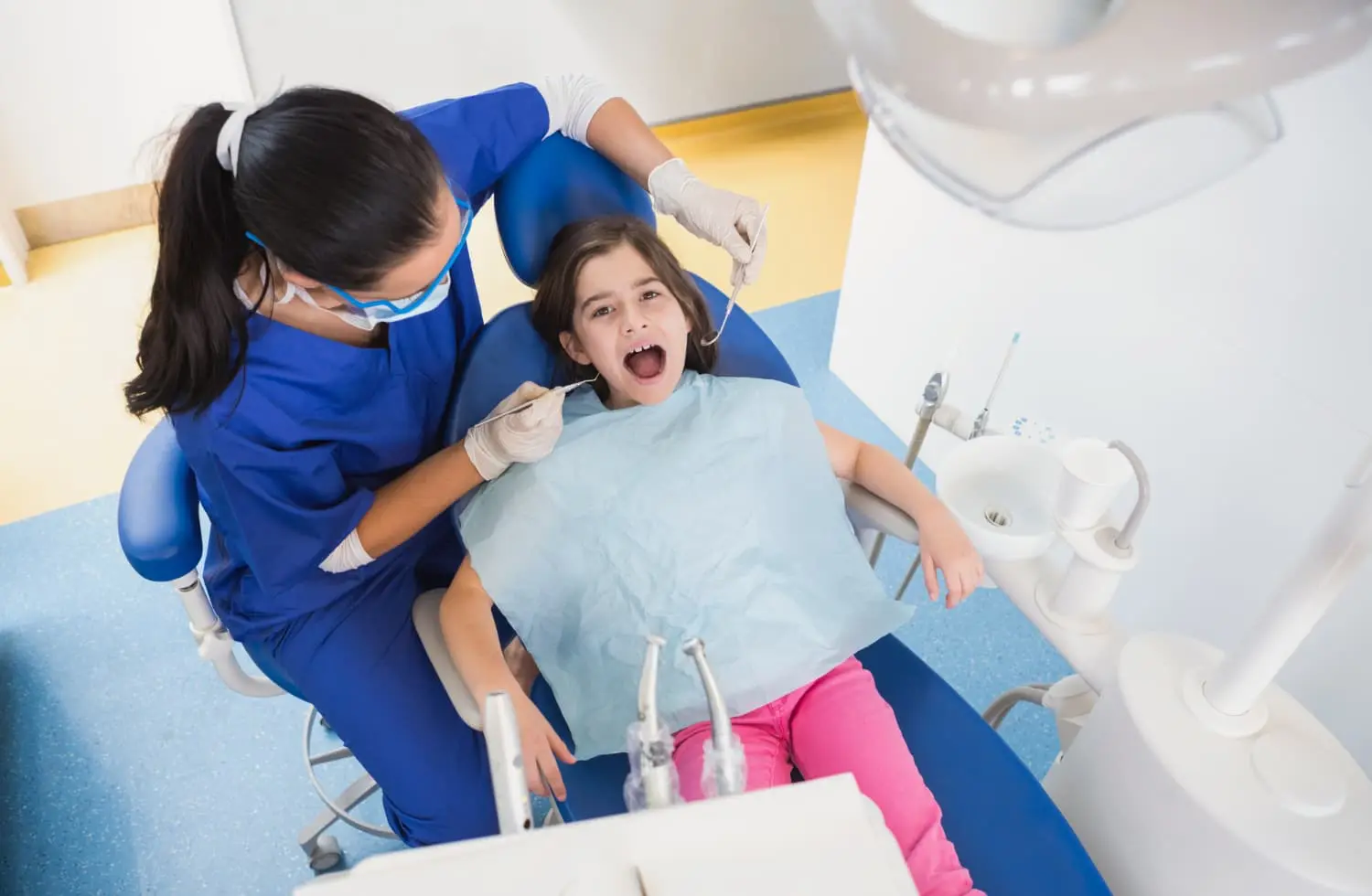 In a well-equipped clinic known for Best Orthodontics, a dentist in blue scrubs examines a young girl sitting in a dental chair, her mouth open and shielded by a protective bib.