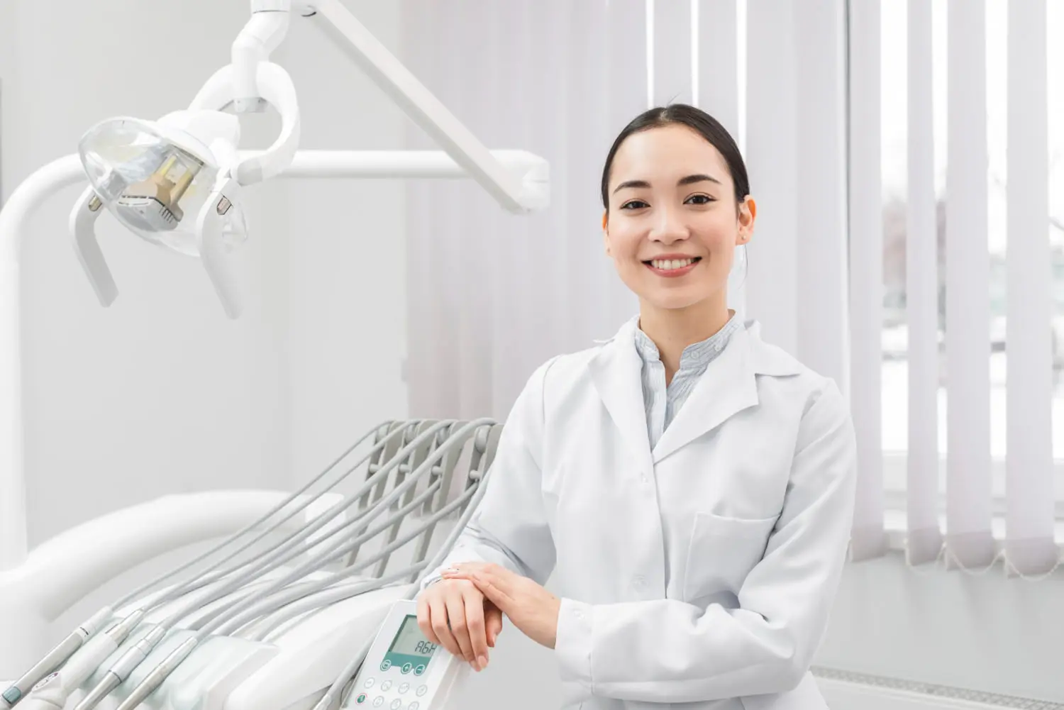 A female dentist in a white coat stands smiling in a dental office, showcasing top-notch orthodontics with dental equipment and a chair in the background.