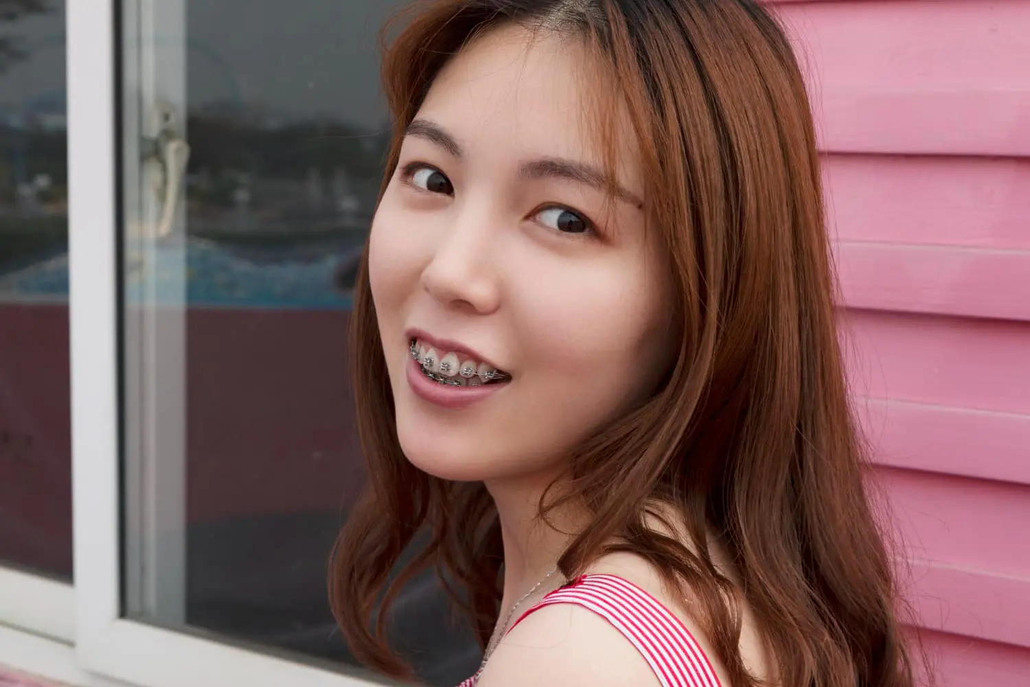A person with long hair and braces, courtesy of Best Orthodontics, smiles brightly in front of a pink wall and window.
