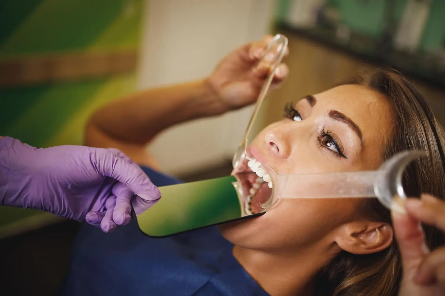 A woman lies back with her mouth open as a dentist in purple gloves, from Best Orthodontics, uses a dental retractor to hold her lips and a mirror to inspect her teeth.