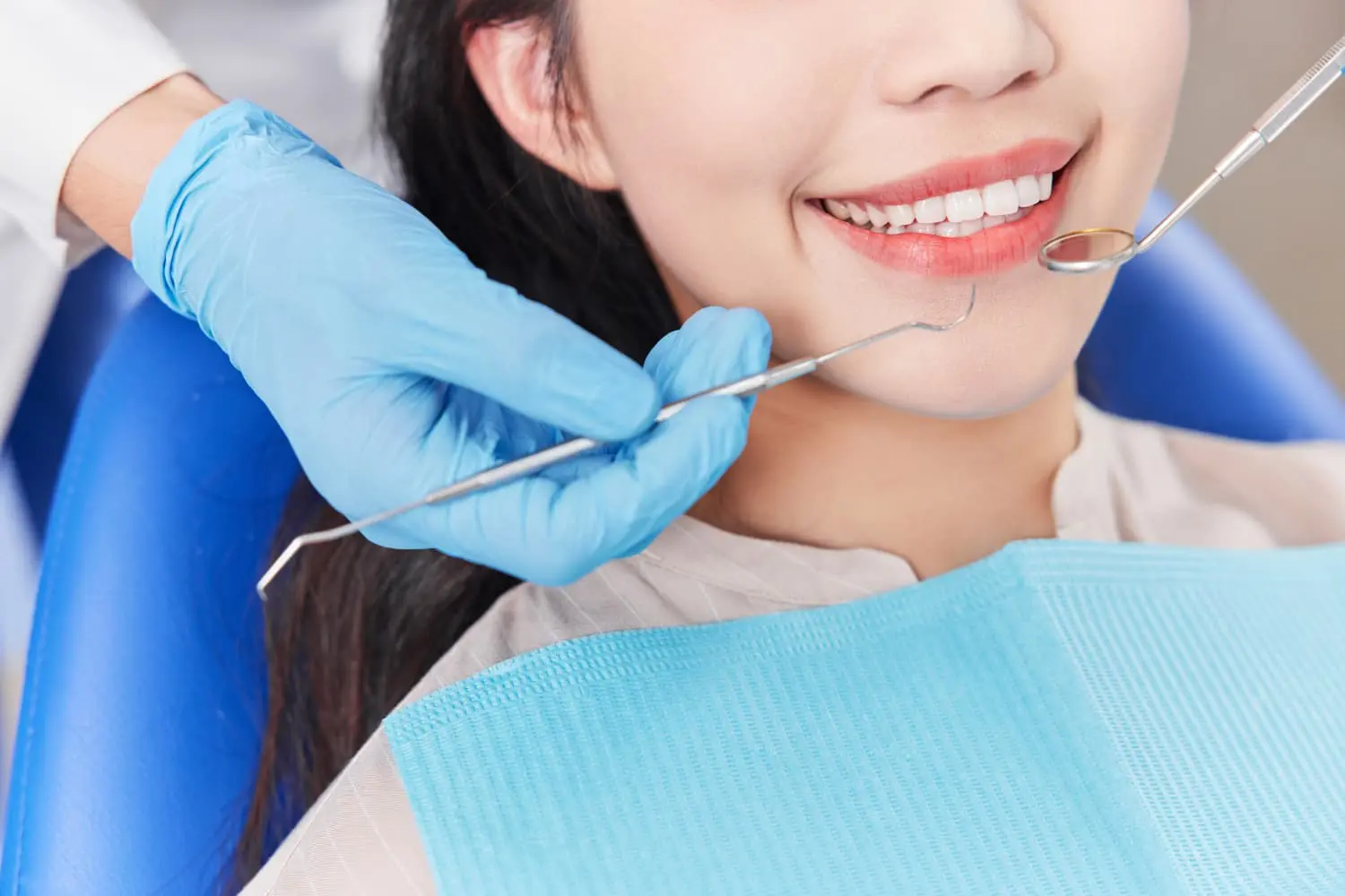 Person receiving dental check-up with a gloved hand holding dental instruments near their mouth; they're smiling while sitting on a dental chair with a blue bib, experiencing the best orthodontics care.