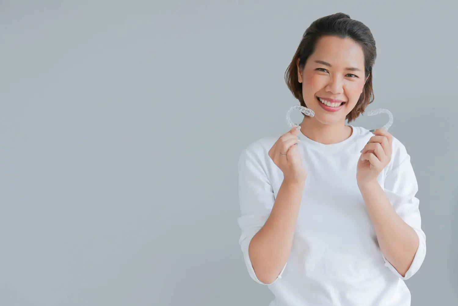 Smiling woman in a white shirt holds two clear dental aligners in front of her, showcasing the expertise of Best Orthodontics, as she stands against a plain gray background.