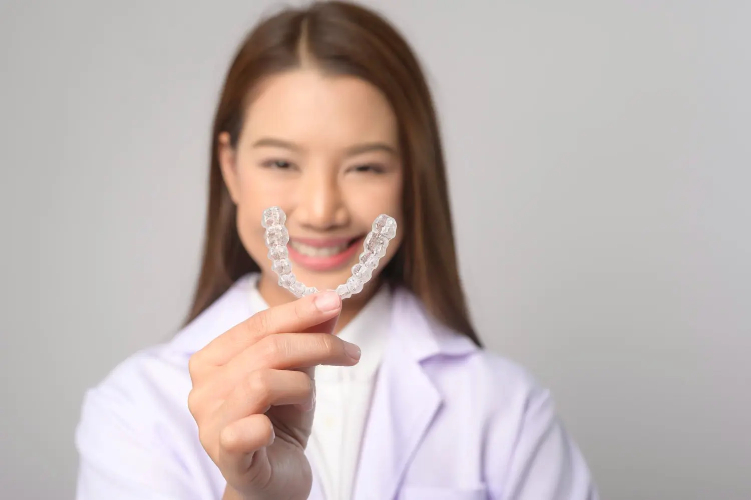 A person in a white coat, representing Best Orthodontics, holds a clear dental aligner towards the camera, smiling against a softly blurred neutral background.