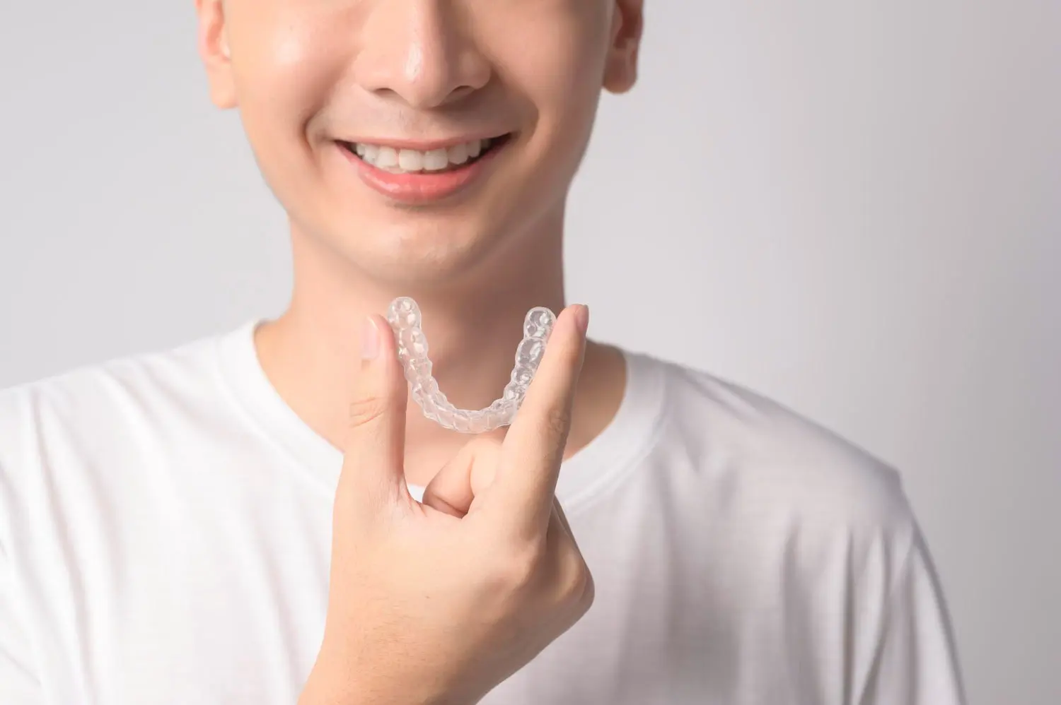 A person, wearing a plain white shirt against a simple background, smiles while holding a clear dental aligner—the hallmark of best orthodontics in modern care.
