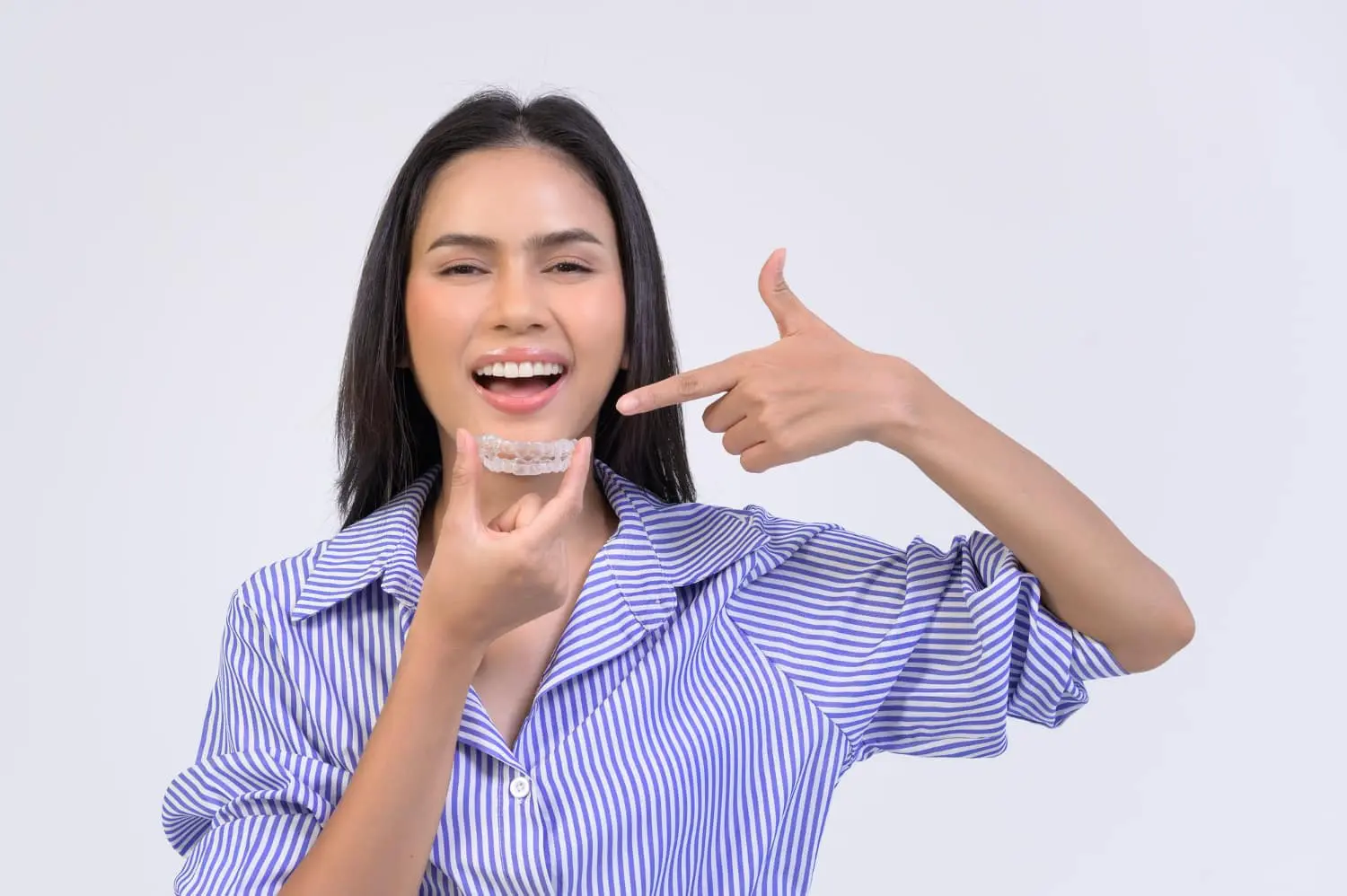 A woman in a striped shirt smiles brightly, showcasing the best orthodontics care as she holds and points to a clear dental aligner.