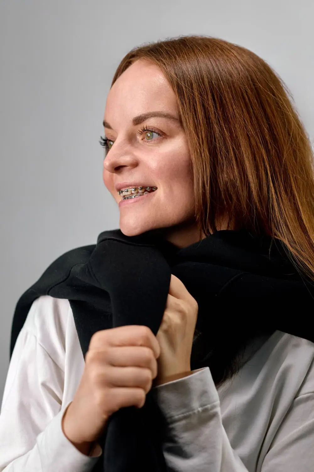 A woman with long brown hair and braces, showcasing the work of Best Orthodontics, wears a white shirt and holds a black jacket. She smiles warmly against a gray background while looking to the side.