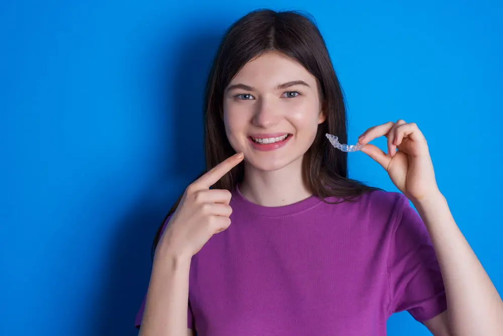 A person smiling and proudly pointing at their teeth, showcasing the effectiveness of Orthoone Orthodontics, holds a clear dental aligner against a vibrant blue background.