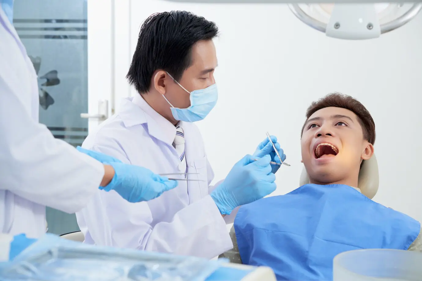 Dentist examines a patient's open mouth while an assistant holds a tray with dental tools. Both wear protective masks and gloves.