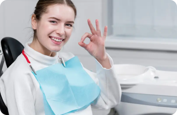 A person in a dental chair wearing a bib smiles and makes an OK gesture.