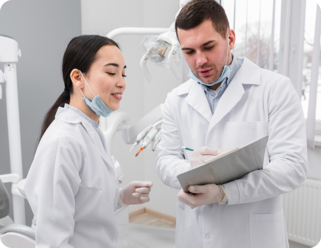 Two dental professionals in white coats and masks discuss in a dental office. One holds a clipboard and dental equipment is in the background.