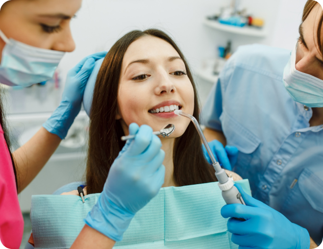A woman is sitting in a dental chair, receiving treatment from two dental professionals wearing masks and gloves.
