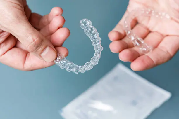 Two hands holding clear Invisalign aligners over a blue surface, with a package in the background in Austin