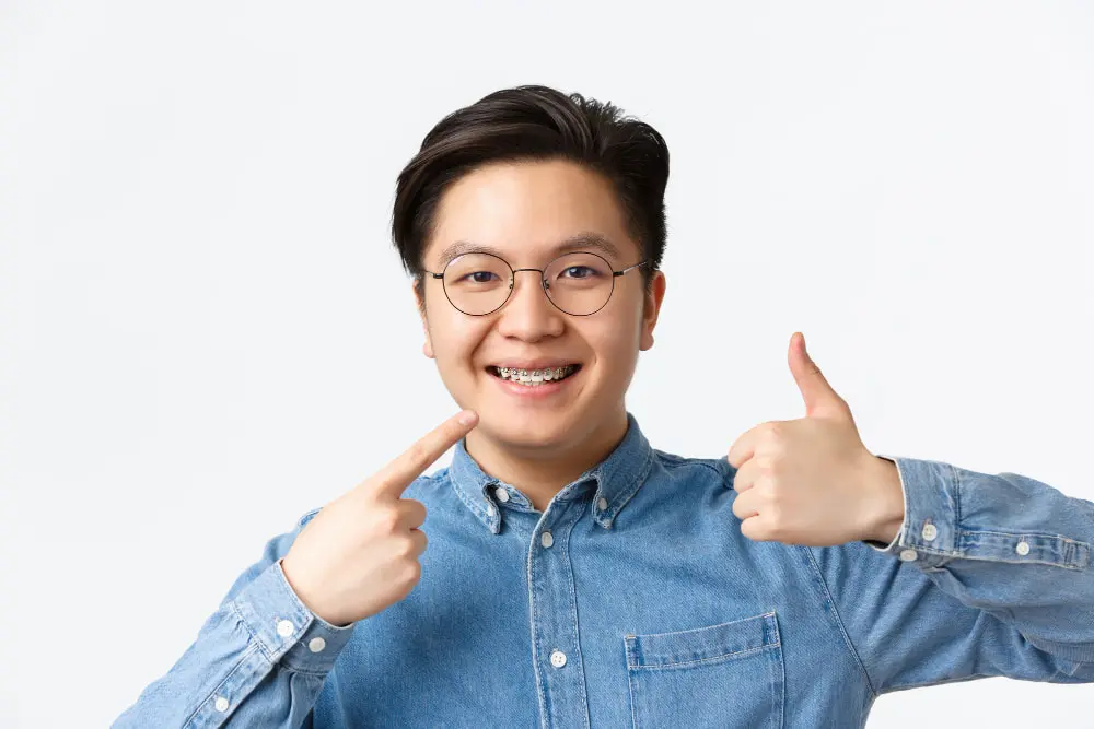 Close-up of satisfied guy, dental clinic client smiling happy and pointing at his dental braces and showing thumbs-up in approval, recommend